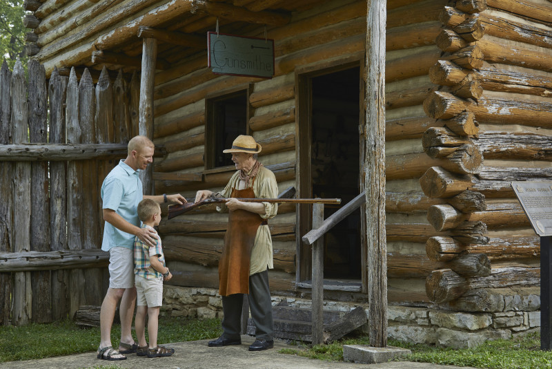 Historic actor with tourists showing old rifle in front of log building.